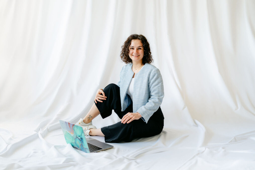 „Belinda, Gründerin von BÉLIE Weddings, sitzt entspannt auf dem Boden in legerer Pose neben ihrem Laptop, lächelt in die Kamera und vermittelt ihre lockere, strukturierte Herangehensweise an individuelle Hochzeitsplanung in Wien und Österreich.“